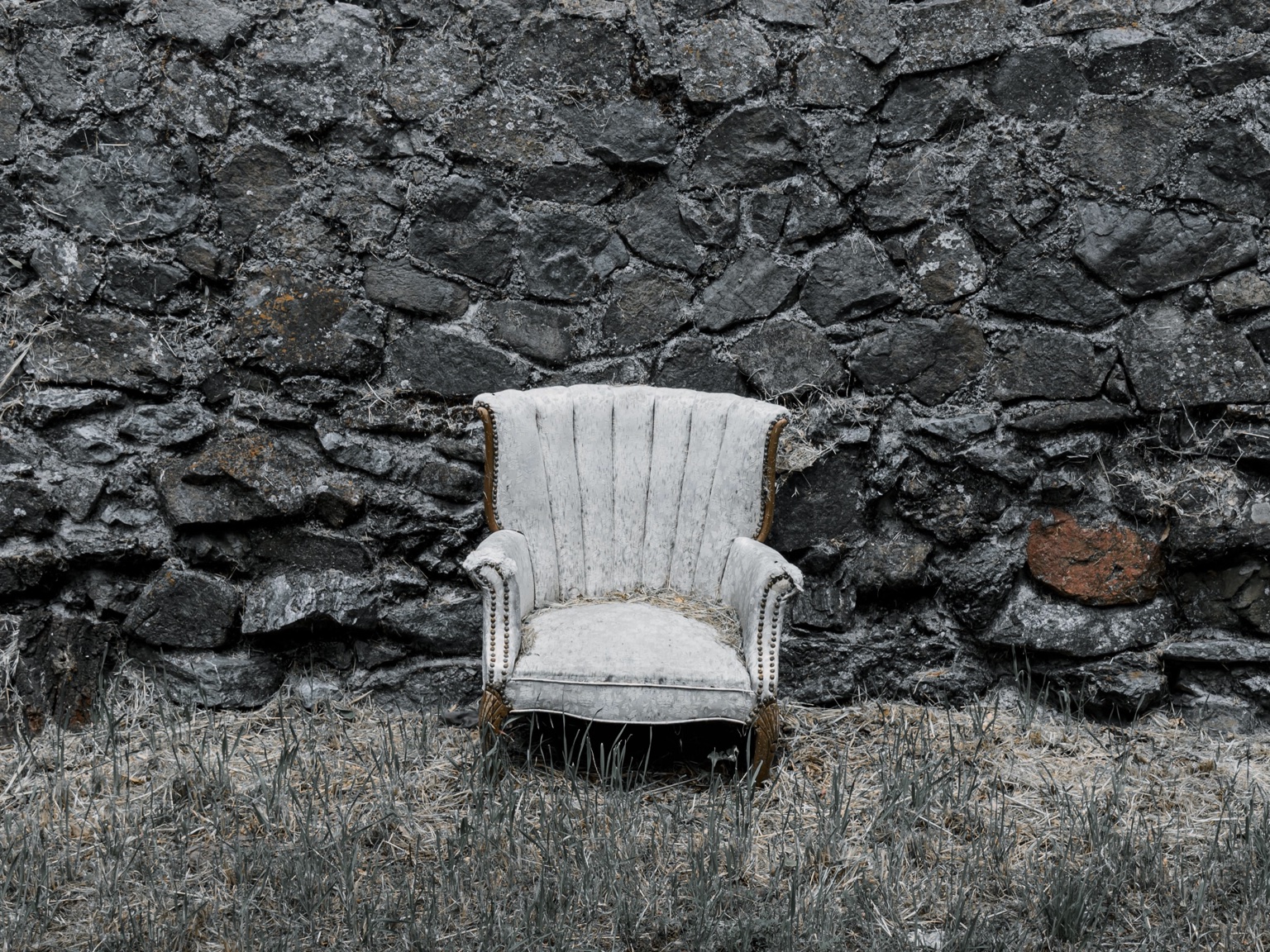 Weathered armchair standing in dry grass before a dark stone wall.